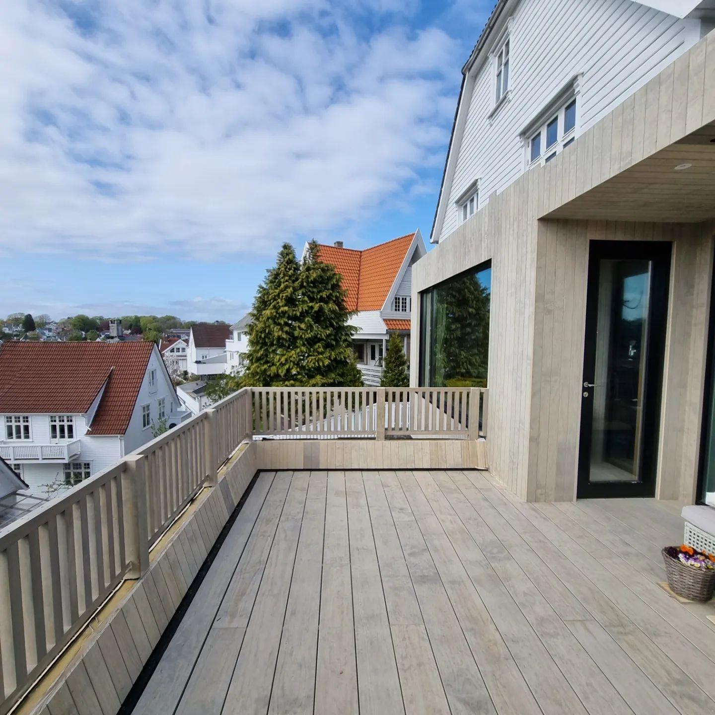 Moderne terrasse med tredekk, utsikt over hus og himmel, omgitt av grønne trær og bygninger.