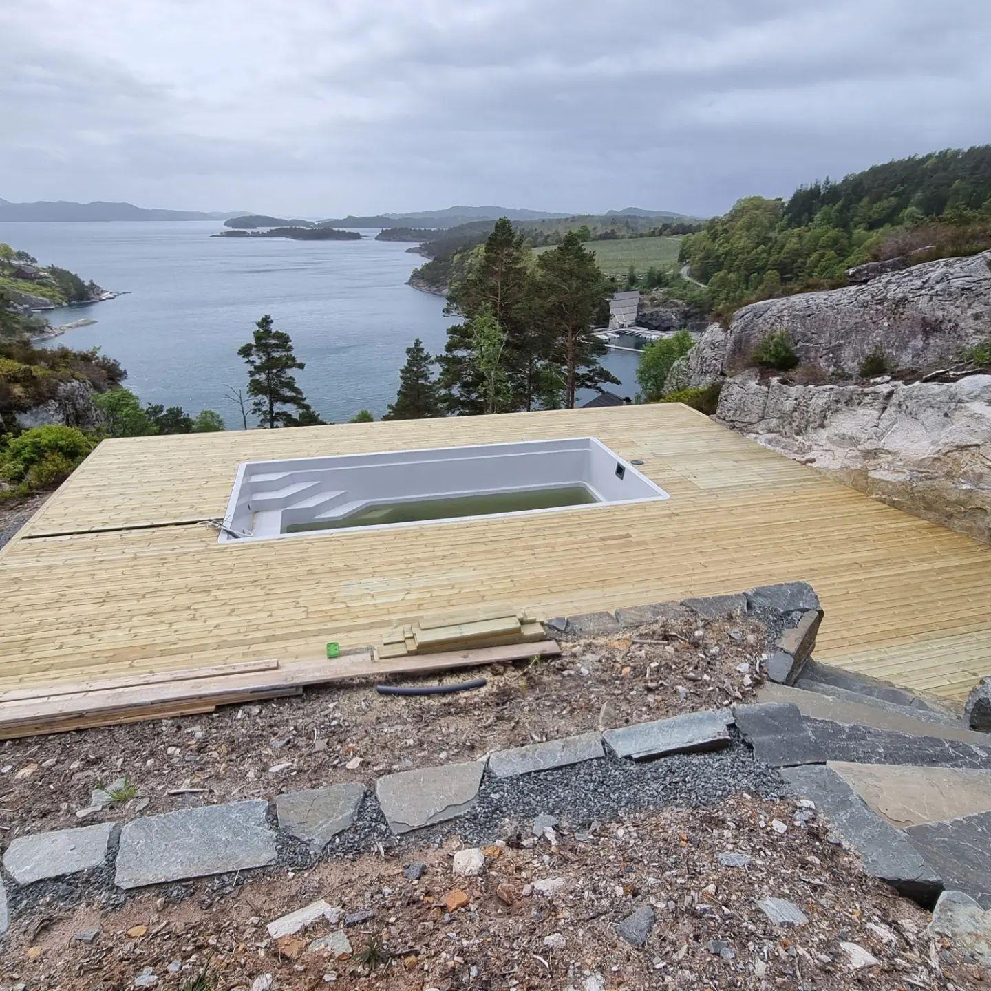 Utsikt over en terrasse med boblebad ved sjøen, omkranset av trær og fjell.