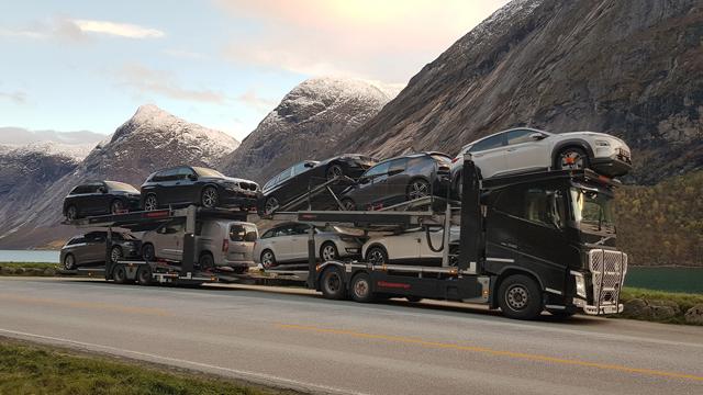 En lastebil med flere biler på tilhenger, parkert langs en vei med fjell og innsjø i bakgrunnen.