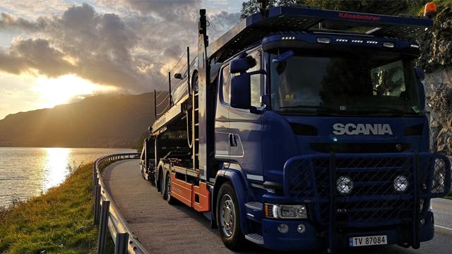 En blå Scania lastebil parkert langs en vei med solnedgang over fjorden.