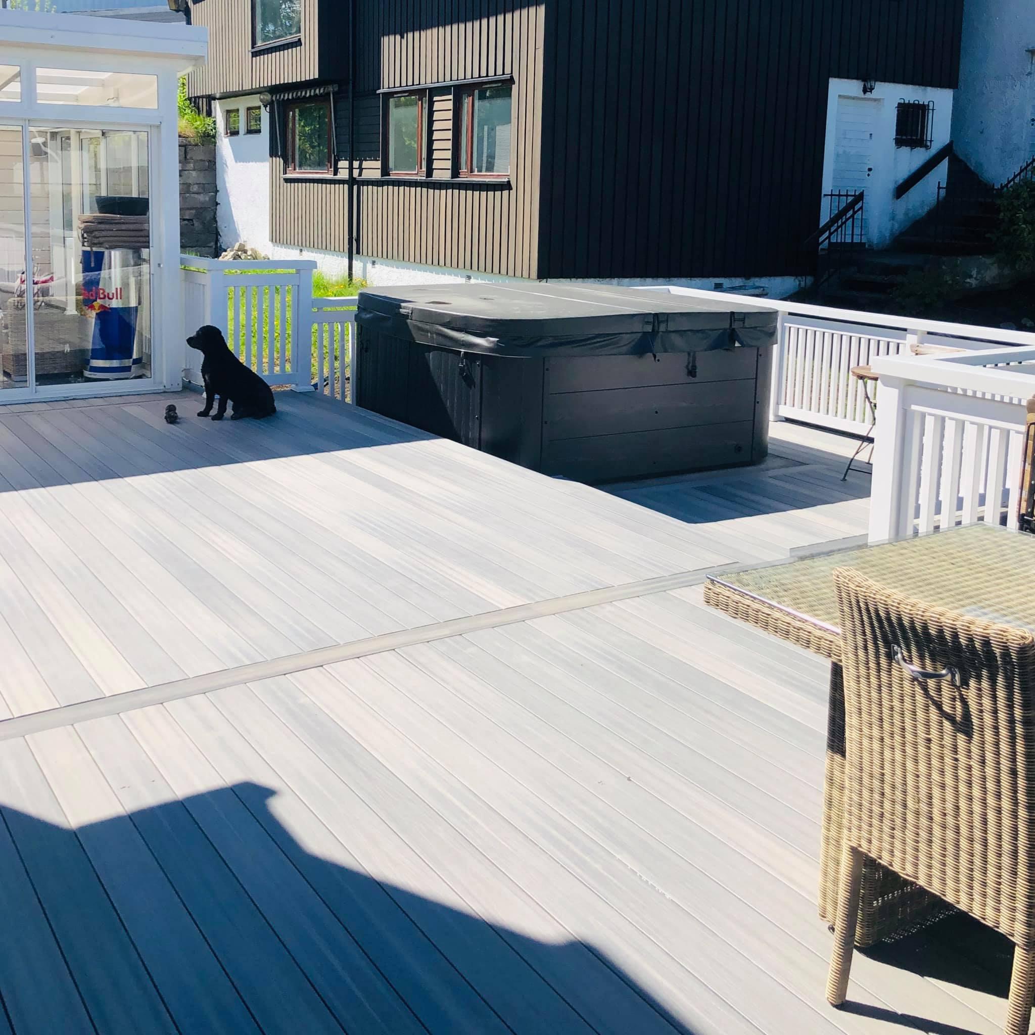 En svart hund sitter på en terrasse med en stor svart boks og en hagebenk i bakgrunnen.