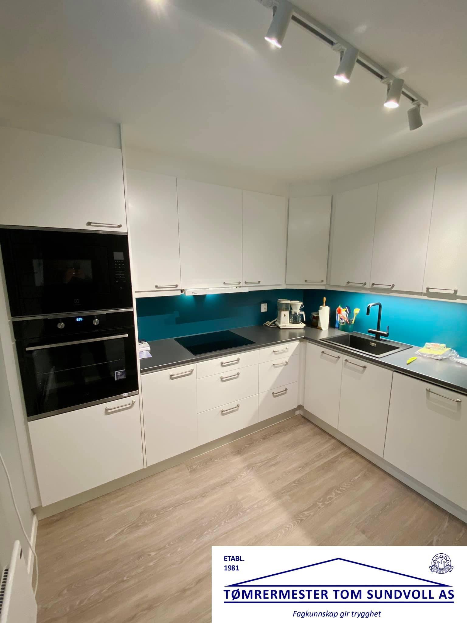 Moderne kjøkken med hvite skap, svart komfyr, og blå veggpanel. God belysning og moderne design.