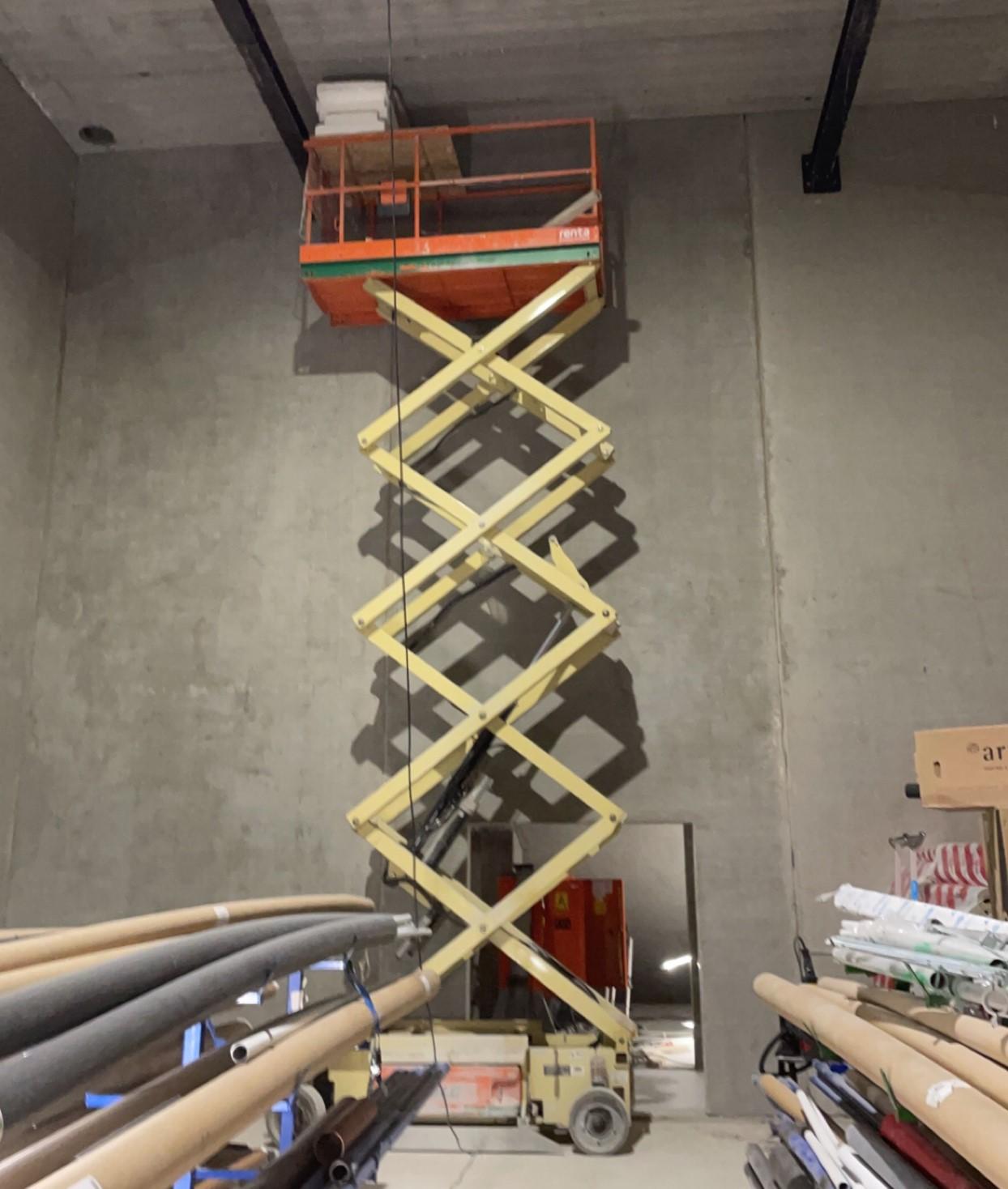 En heis med plattform står oppe i et rom med betongvegger, omringet av rør og byggeutstyr.