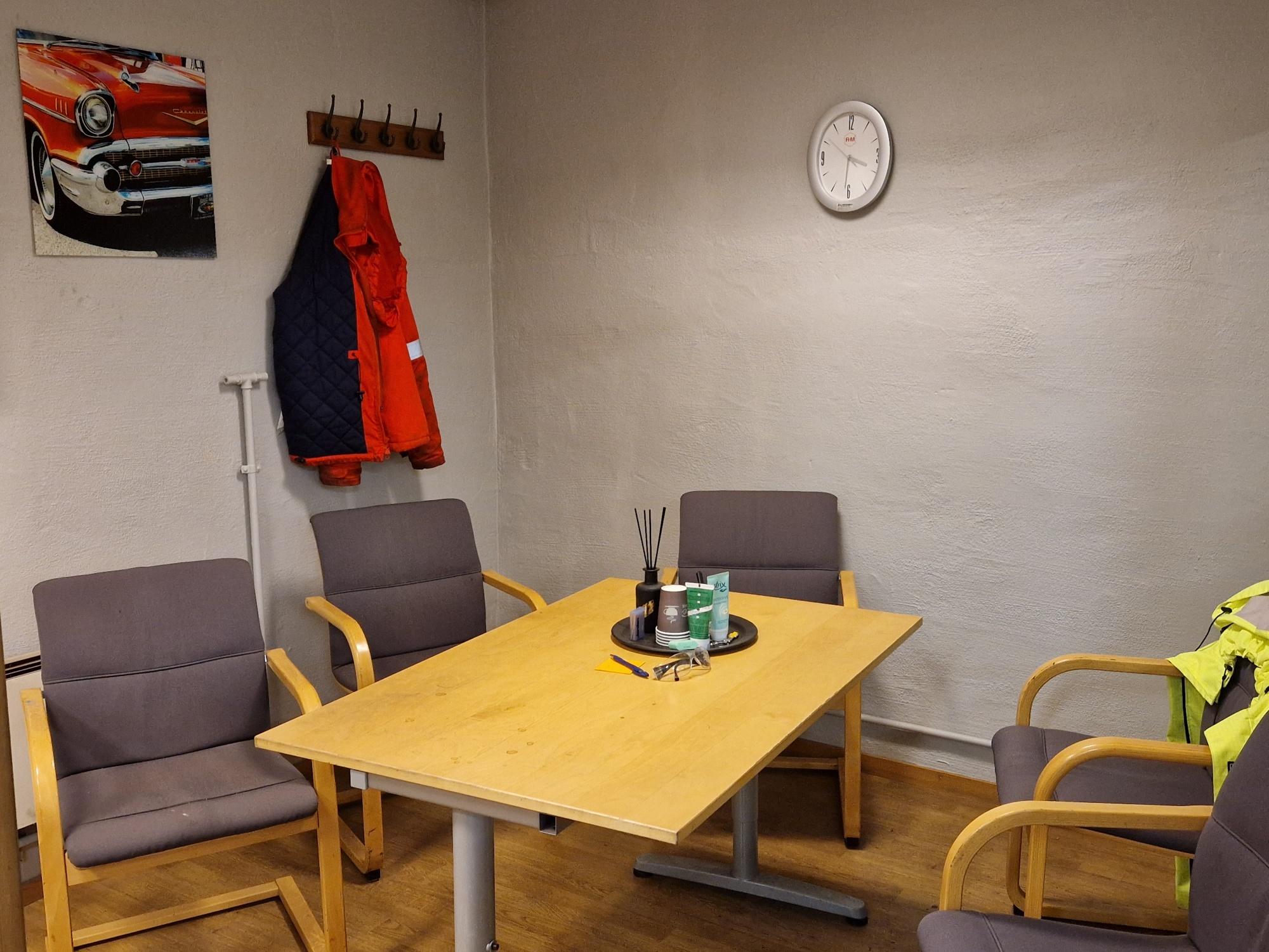 Møterom med fire stoler, et bord, en klokke på veggen og en rød jakke hengende på knaggen.