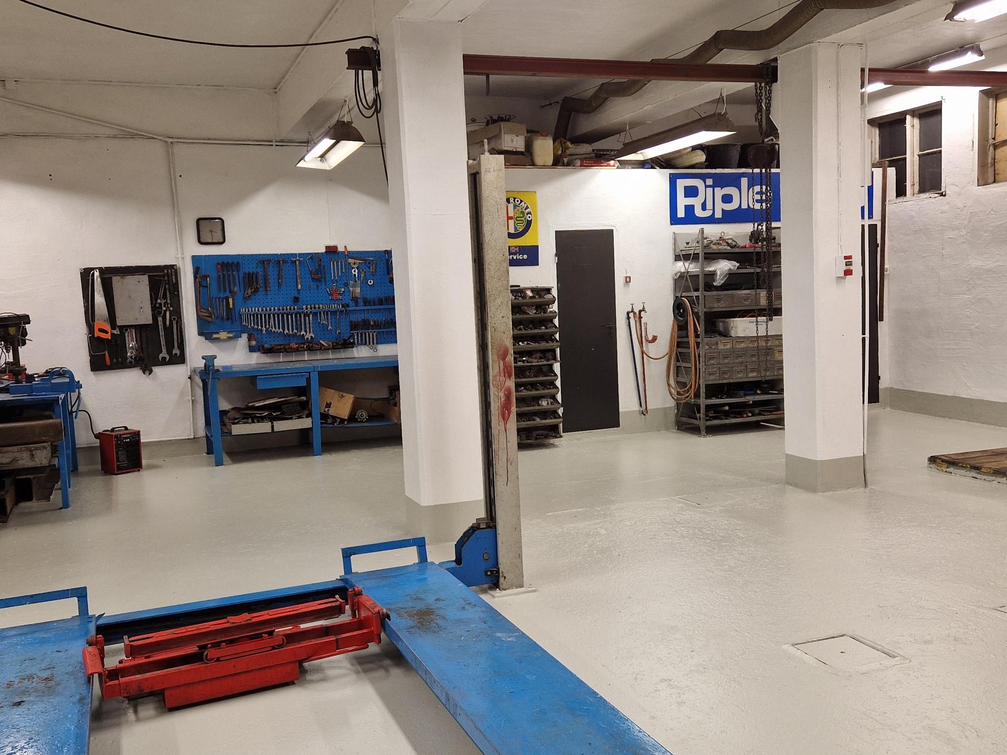 Verksted med blå arbeidsbenker, verktøy på veggen og en heis i midten av rommet.
