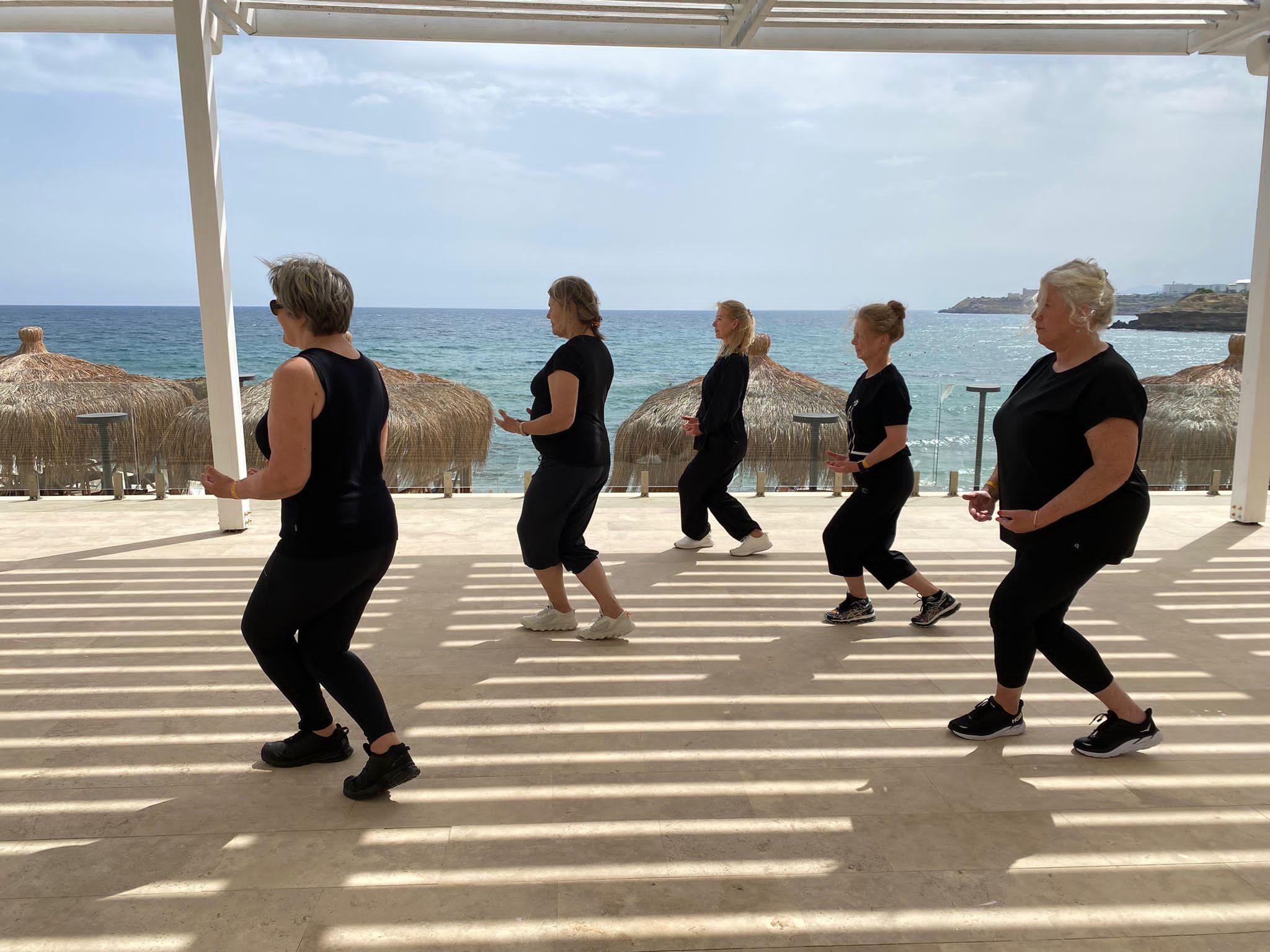 Fem kvinner danser i takt på en strandpromenade med havet i bakgrunnen. Sollys skaper striper på bakken.