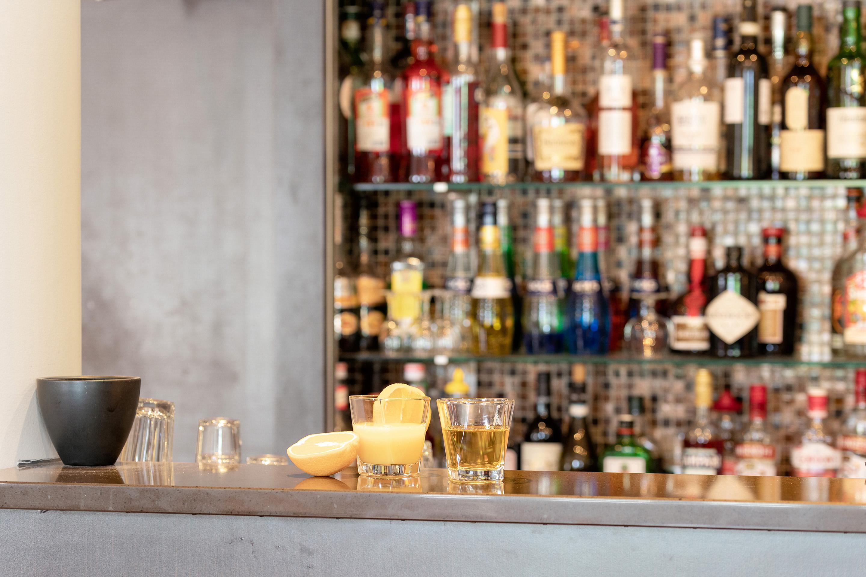 En bar med et variert utvalg av flasker, to glass med drinker og en skål med sitron på disken.