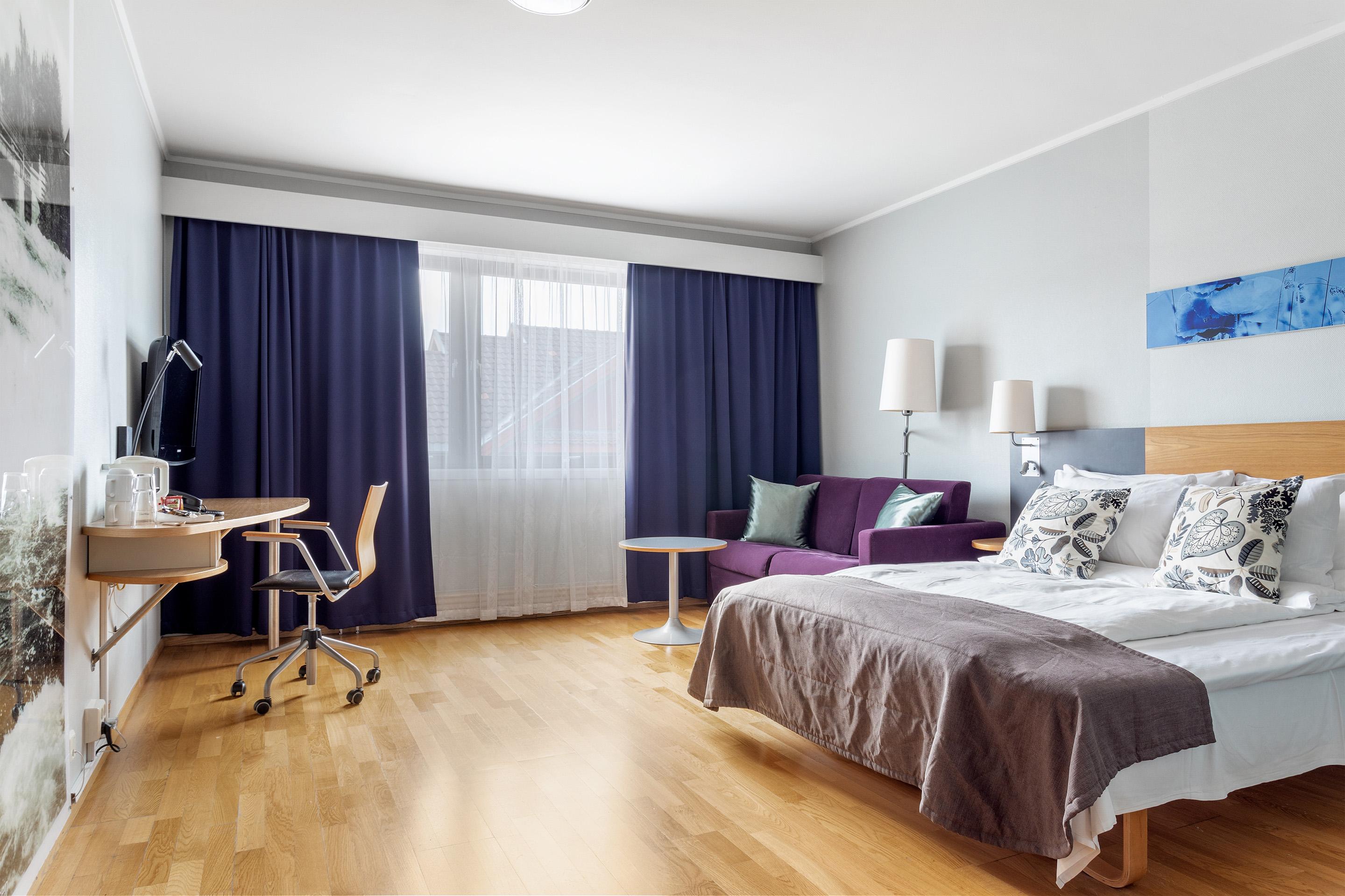 Moderne hotellrom med dobbeltseng, skrivebord, sofa og vinduer med gardiner i blått og grått.
