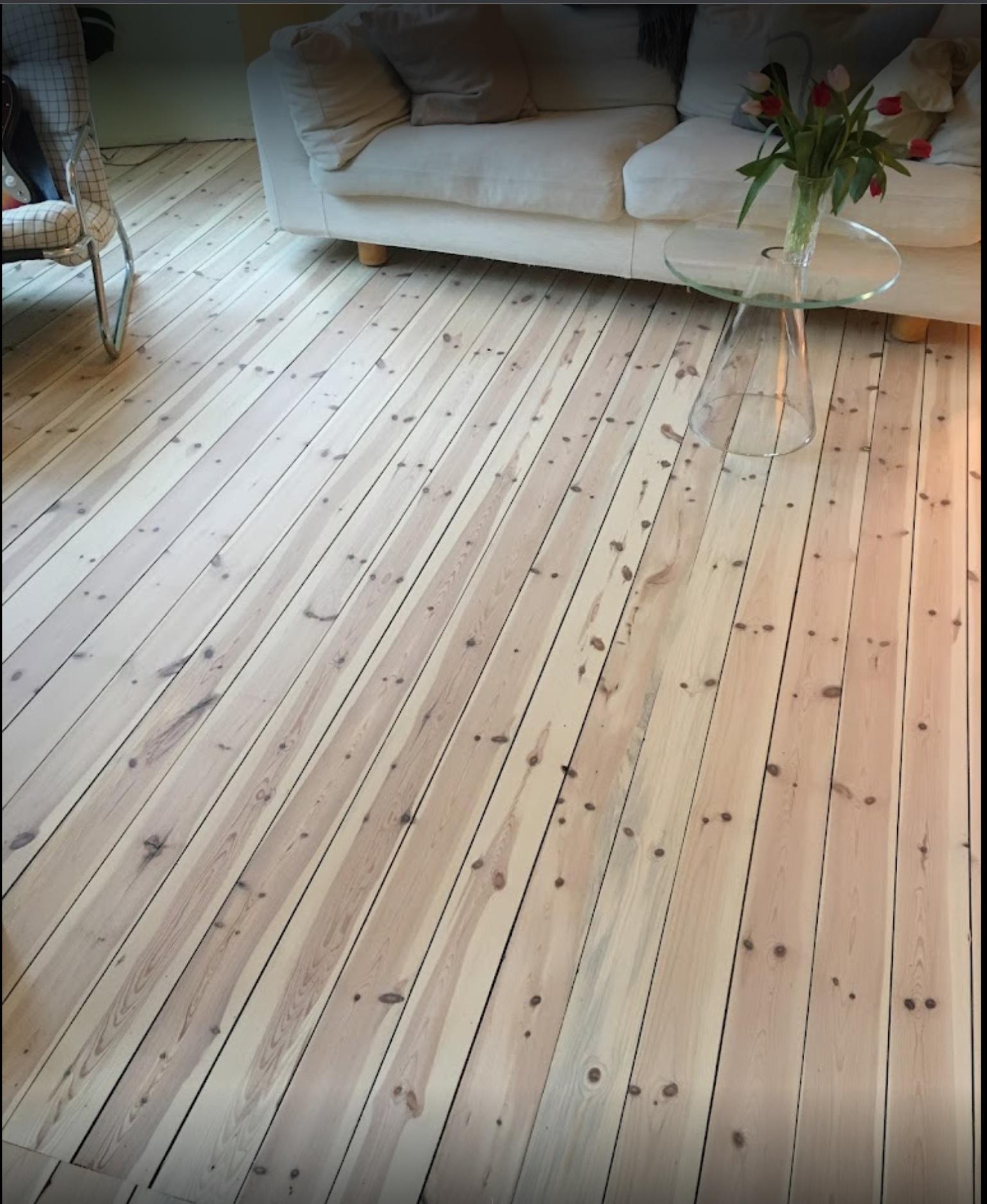 Lyst tregulv med synlige kvister, sofa i bakgrunnen og et glassbord med blomster.