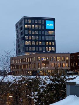 Moderne kontorbygg med Aider-logo, beliggende i en snødekt bylandskap under grå himmel.
