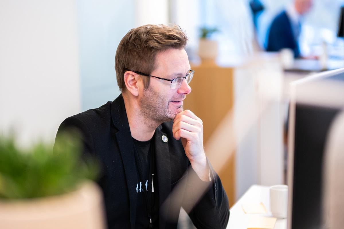 En mann med briller sitter ved en datamaskin og smiler, med en plante i forgrunnen.