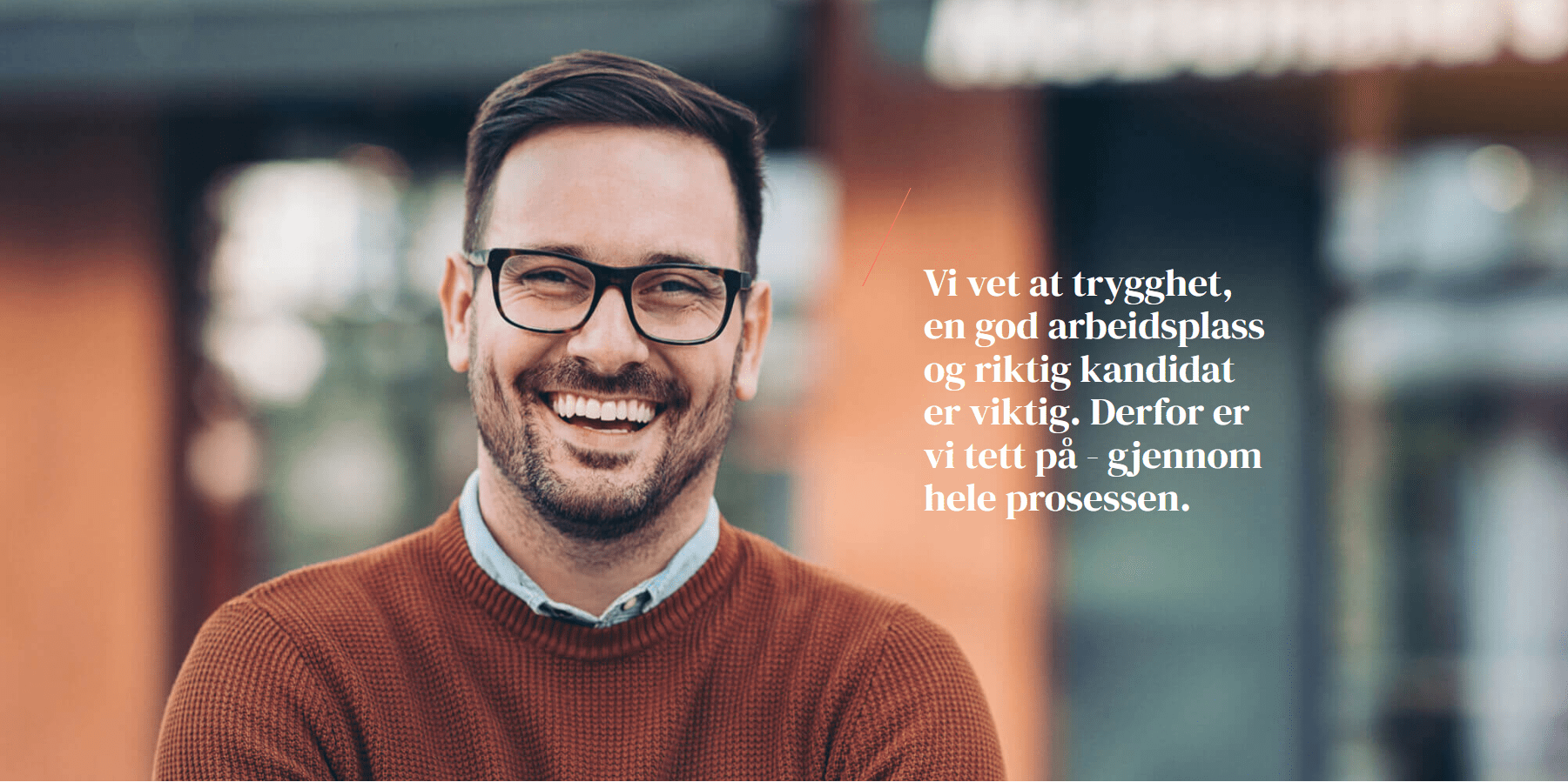 Smilende mann med briller iført brun genser, står foran en bygning med tekst om arbeidsplasser.