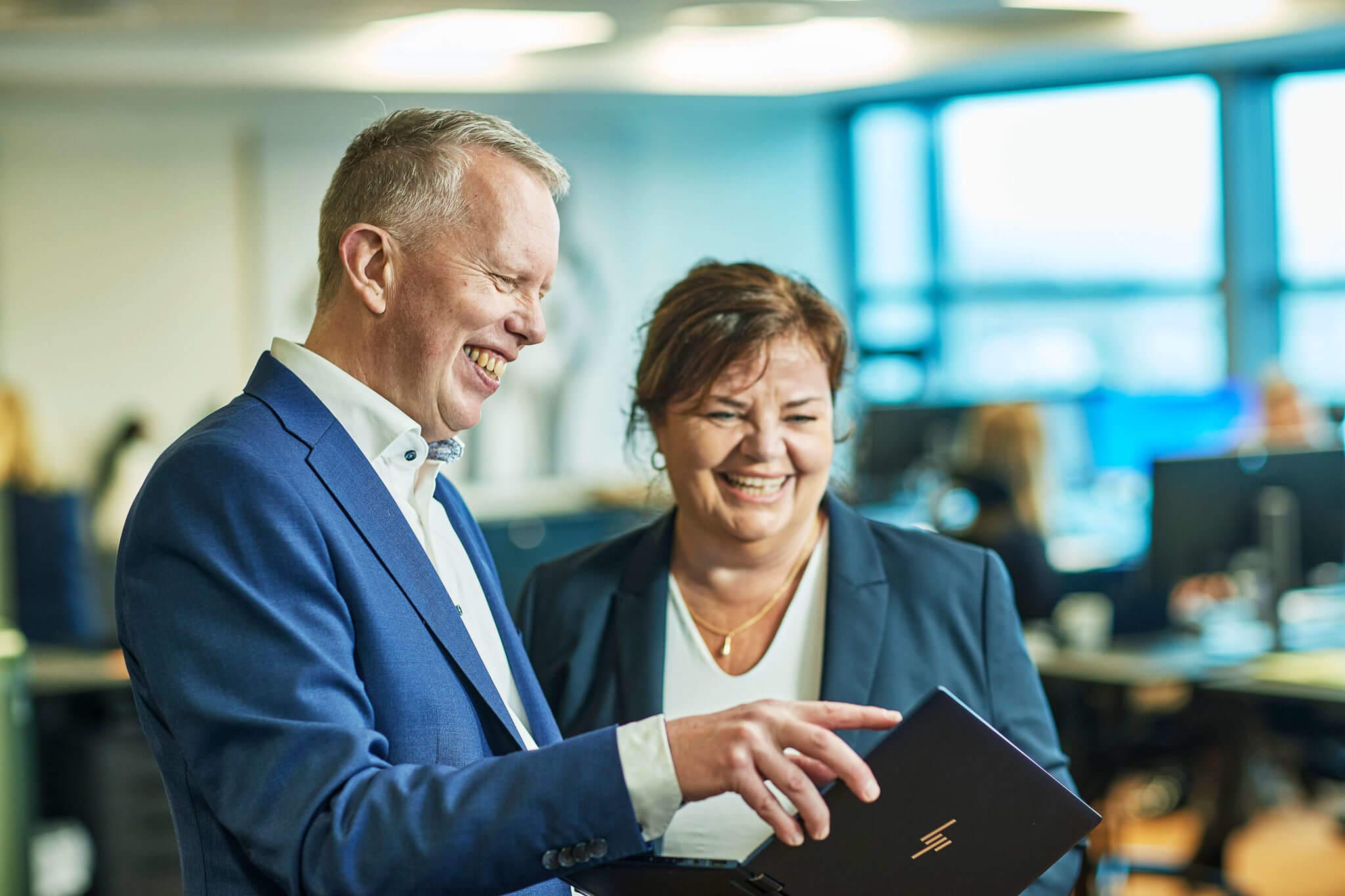 To personer i kontormiljø, smilende og ser på en mappe sammen, med datamaskiner i bakgrunnen.
