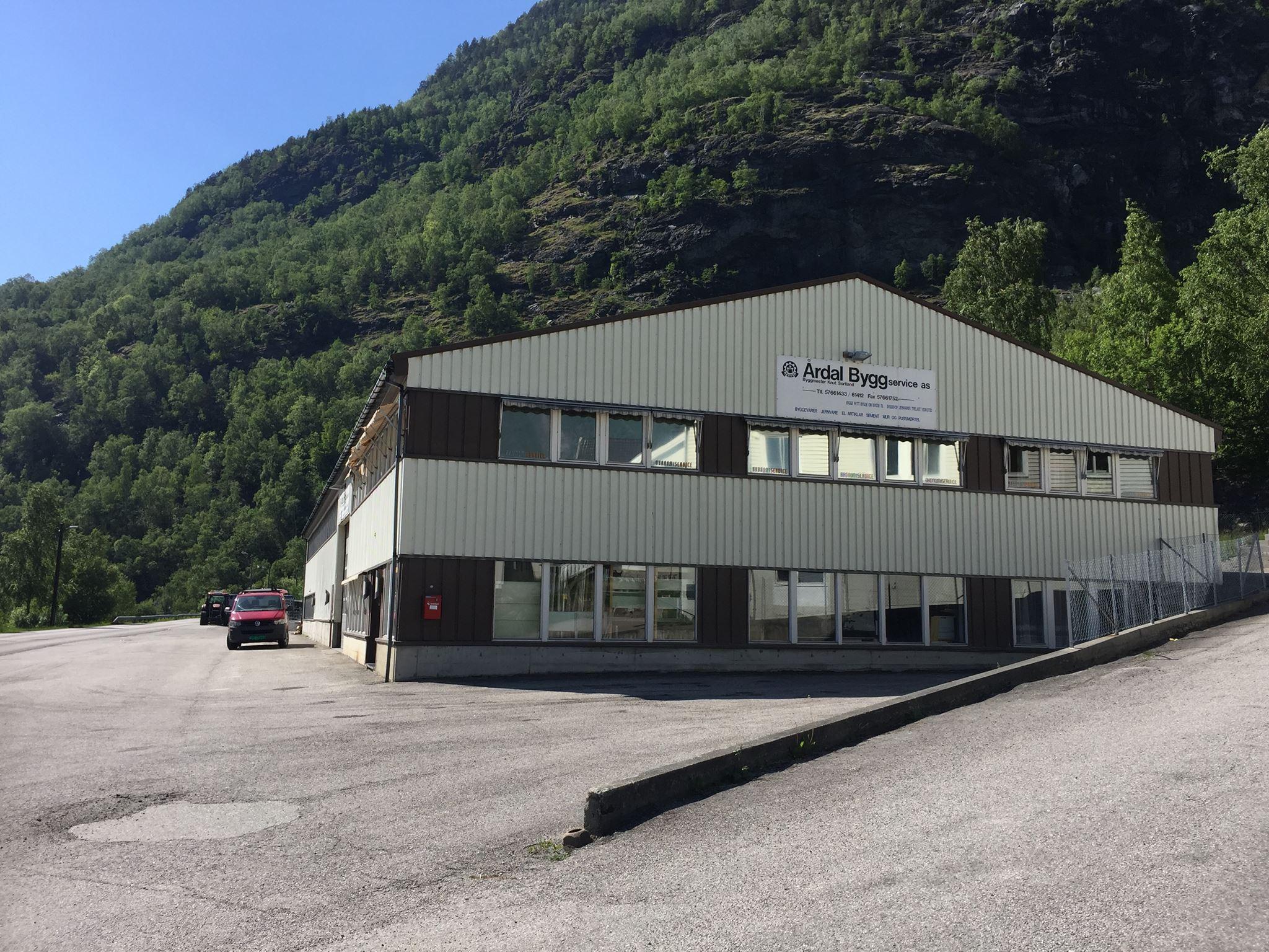 Moderne bygning med flere vinduer, beliggende ved foten av en grønn fjellside. Parkeringsplass foran bygningen.