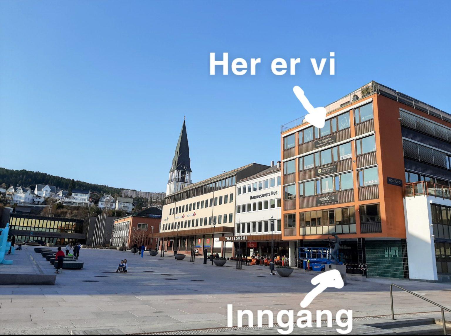 Moderne bygninger med inngang og kirketårn i bakgrunnen, på en solfylt dag i byen.