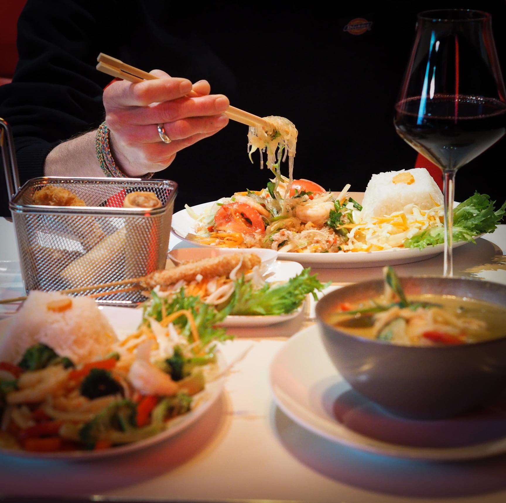 En person som spiser nudler med spisepinner, omgitt av asiatisk mat, ris, salat og et glass rødvin.