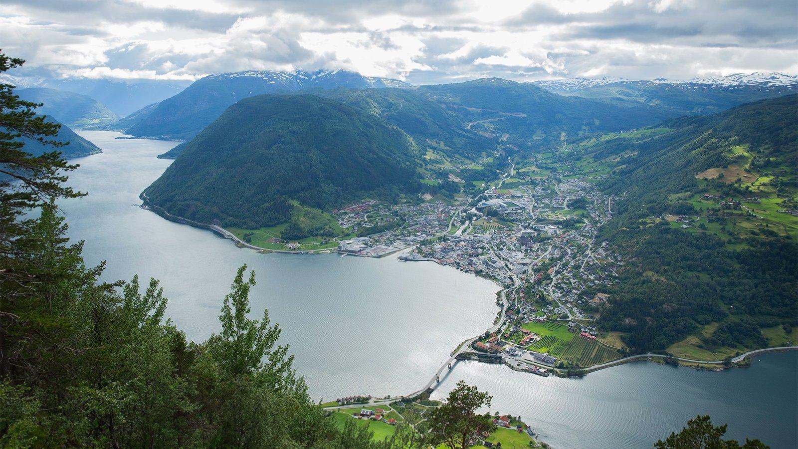 En panoramautsikt over en fjord med en liten by ved vannkanten, omringet av fjell og grønne åser.