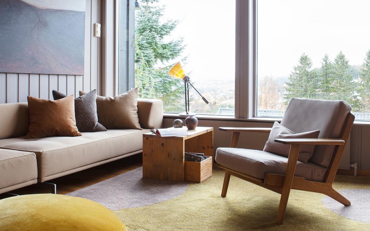 Lyst og moderne stue med beige sofa, trebord, stilig stol og store vinduer som gir utsikt til trær utenfor.