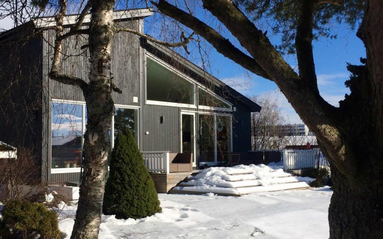 Moderne hytte med mørk fasade, snødekt hage og trær i forgrunnen, under en klar blå himmel.