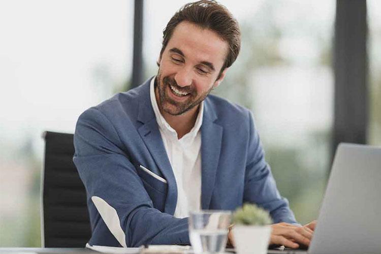 En smilende mann i blå dressjakke sitter ved et skrivebord med en laptop og et glass vann.