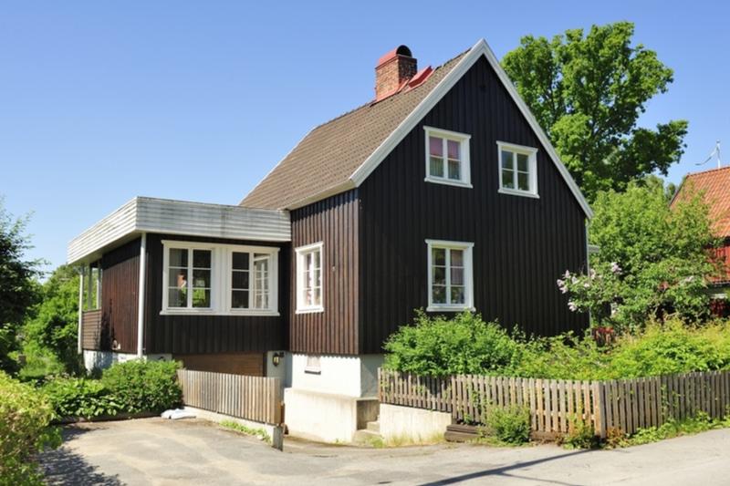 En koselig, svart trehus med hvite vinduer og en liten hage, omgitt av et gjerde på en solfylt dag.