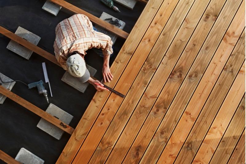 En håndverker legger planker på en treterrasse, med verktøy og materialer rundt seg.