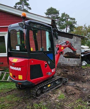 Rød gravemaskin står på en byggeplass ved en rød bygning med grå murvegg.