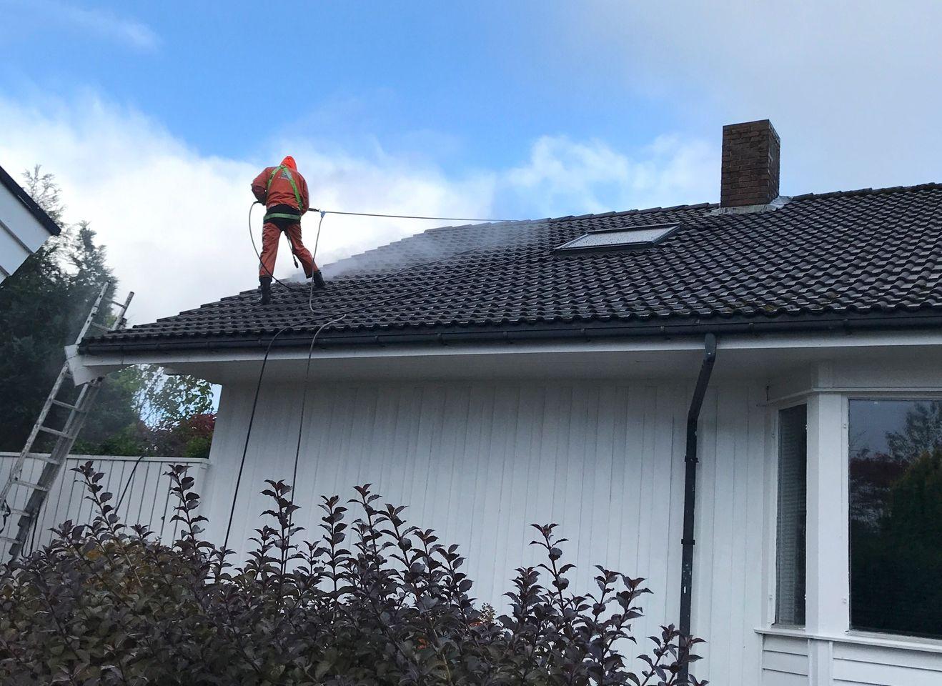 En person i arbeidsoveralls som vasker taket på et hus med høytrykkspyler.