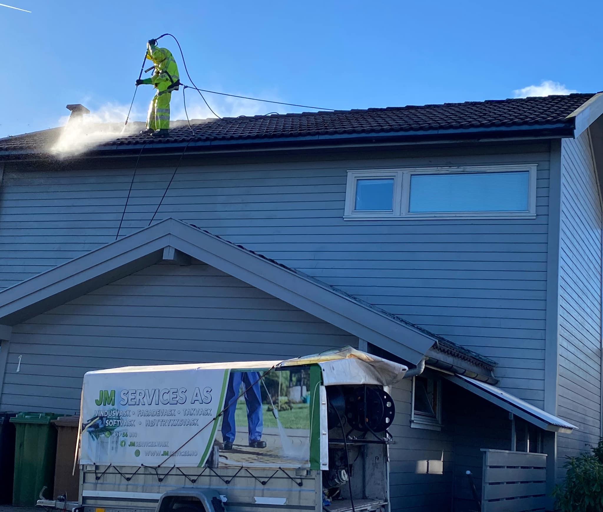 En person i arbeidsklær vasker taket på et hus med høytrykkspyler, med en trailer parkert foran.