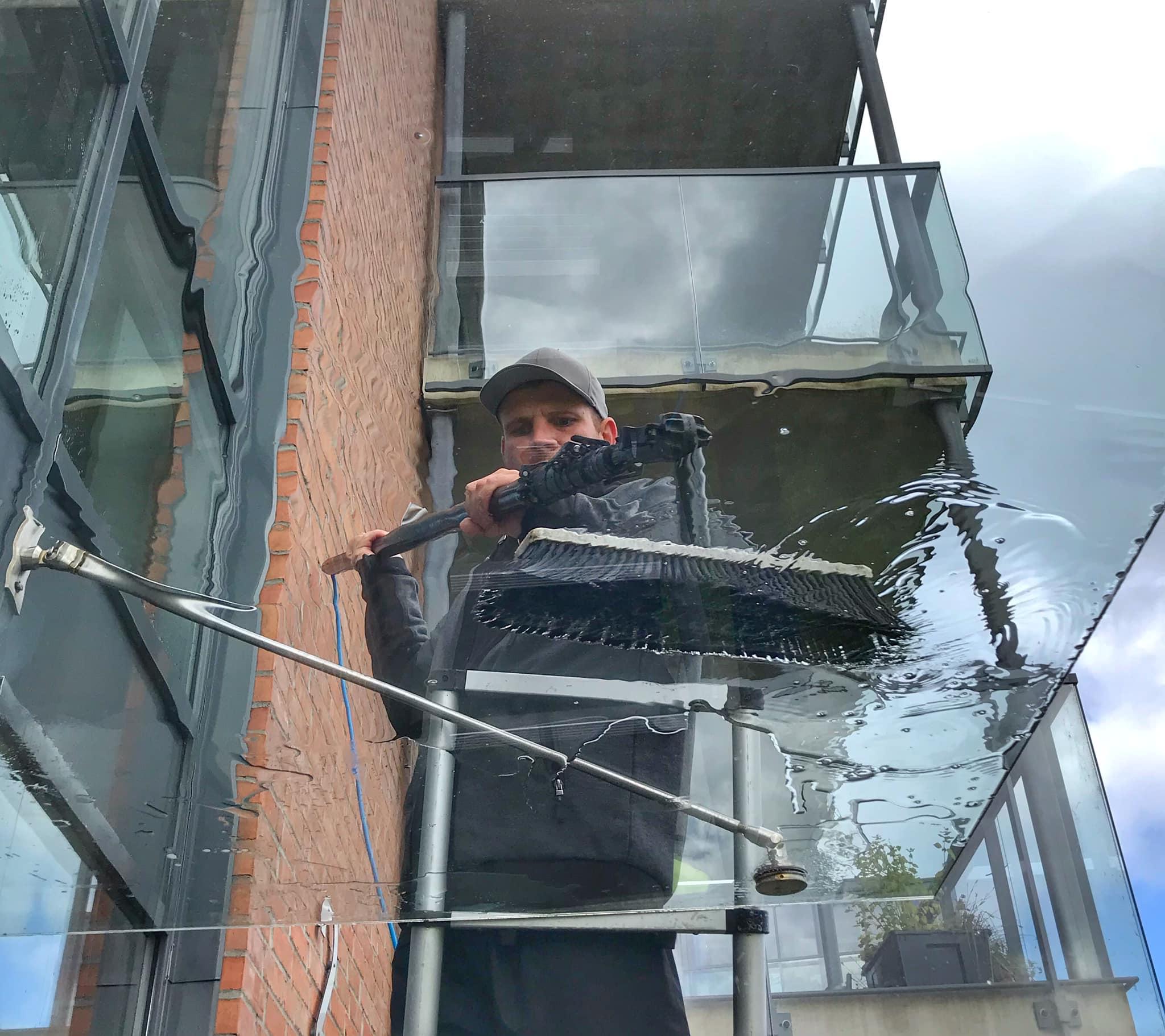 En mann vasker vinduer på en balkong med en lang børste, med bygningen i bakgrunnen.