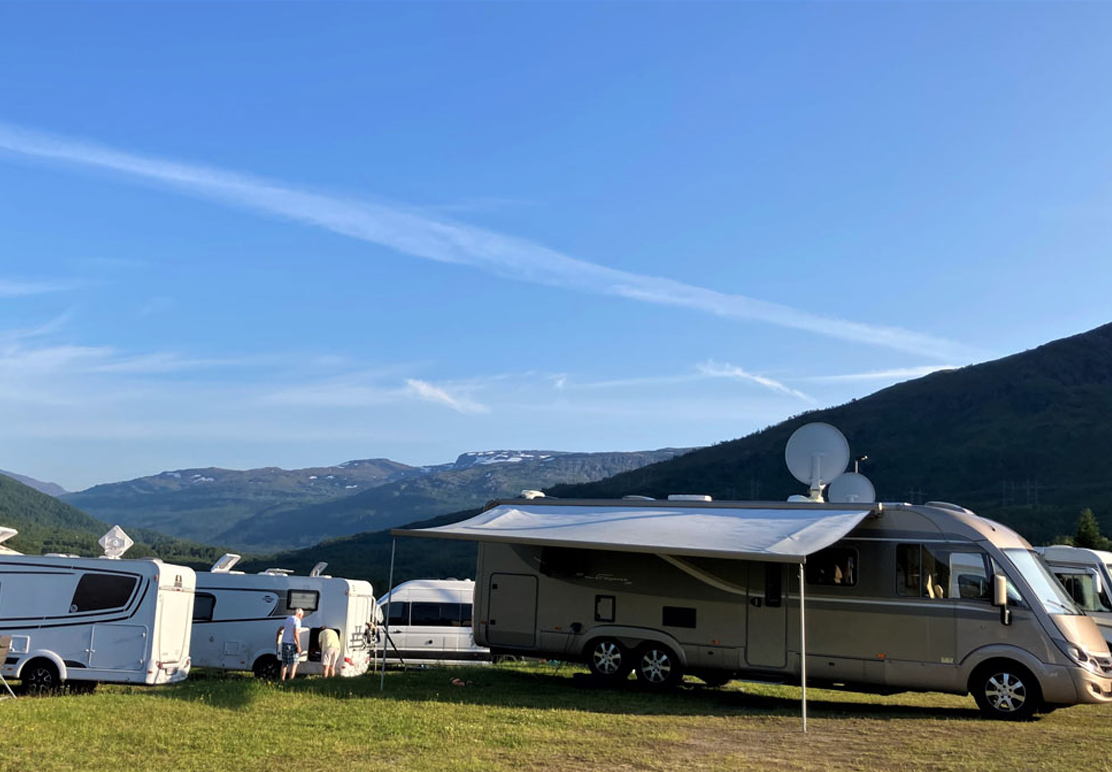 Campingvogn og bobil på en campingplass med fjell i bakgrunnen og blå himmel.