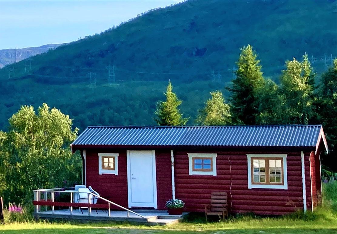 Rødt hytte med hvit dør og vinduer, omgitt av grønne trær og fjell i bakgrunnen.