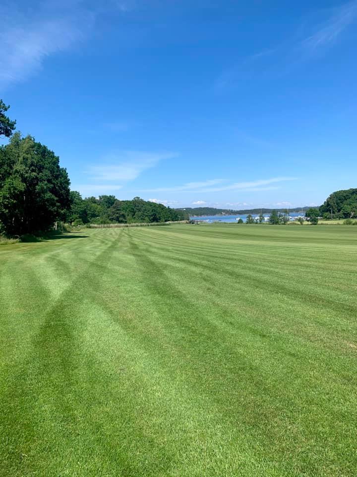En grønn golfbane med striper i gresset, omgitt av trær og en innsjø i bakgrunnen under en klar blå himmel.