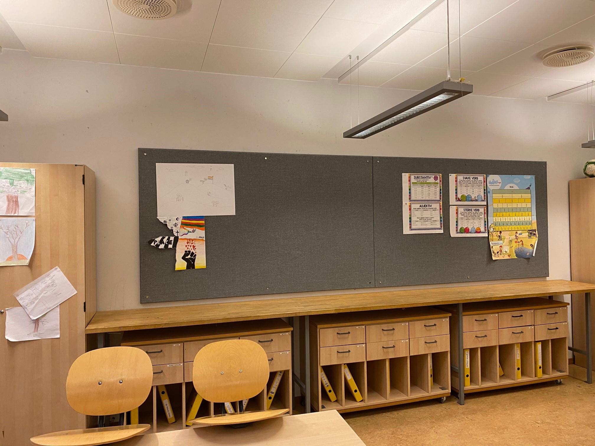 Klasserom med en stor tavle, trevegger, og to trestoler foran en lang benk med oppbevaringsskap.