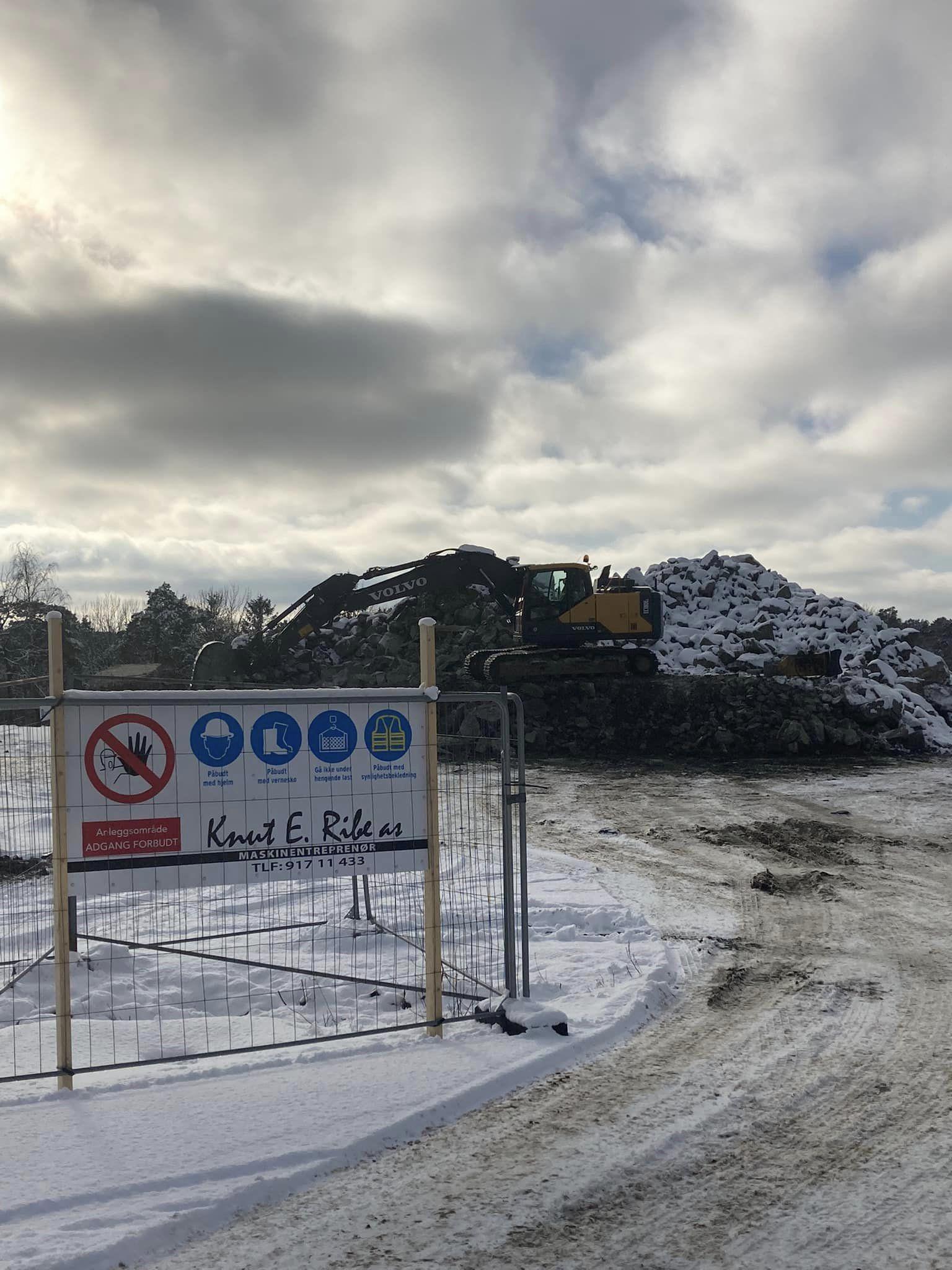 En gravemaskin arbeider på en snødekt byggeplass med en stor haug av jord og et skilt som viser sikkerhetsregler.