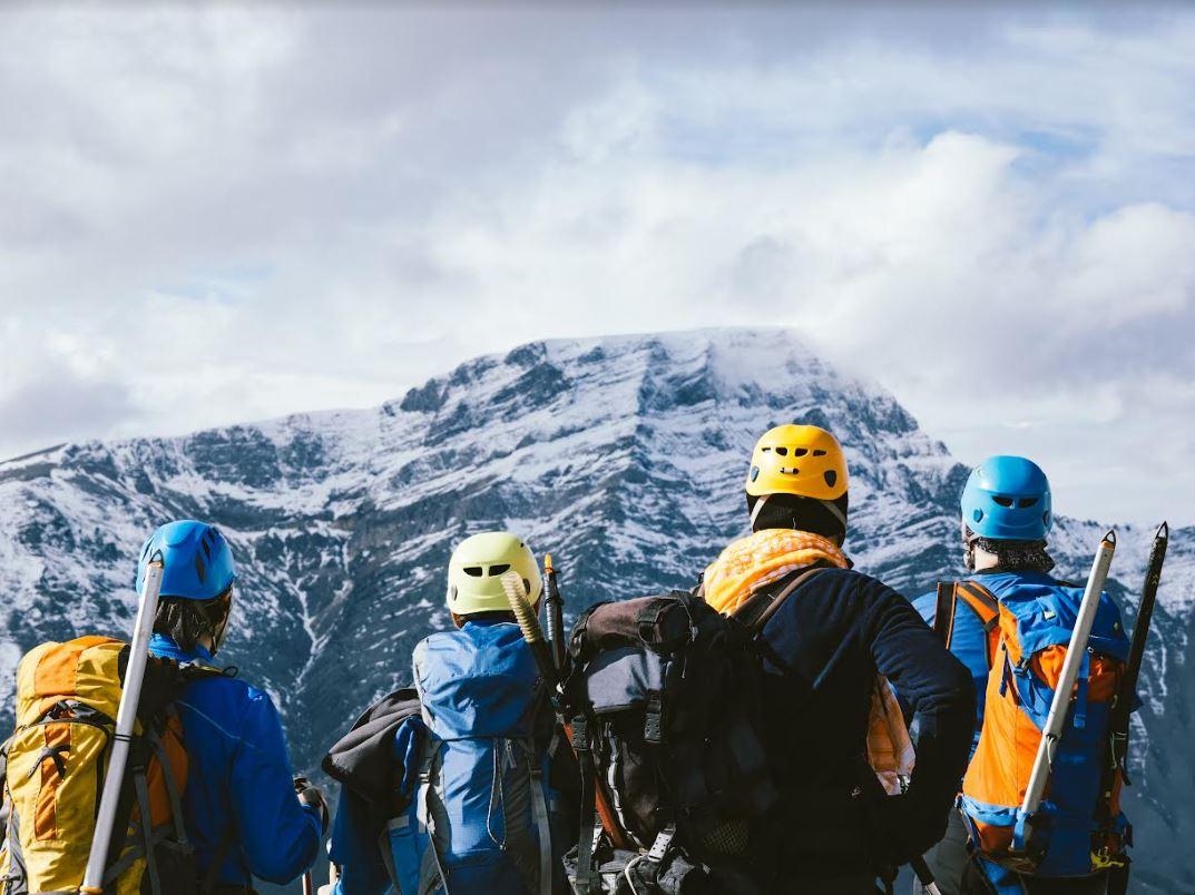 Fire fjellklatrere med ryggsekker og ski ser mot en snødekt fjelltopp under en skyet himmel.