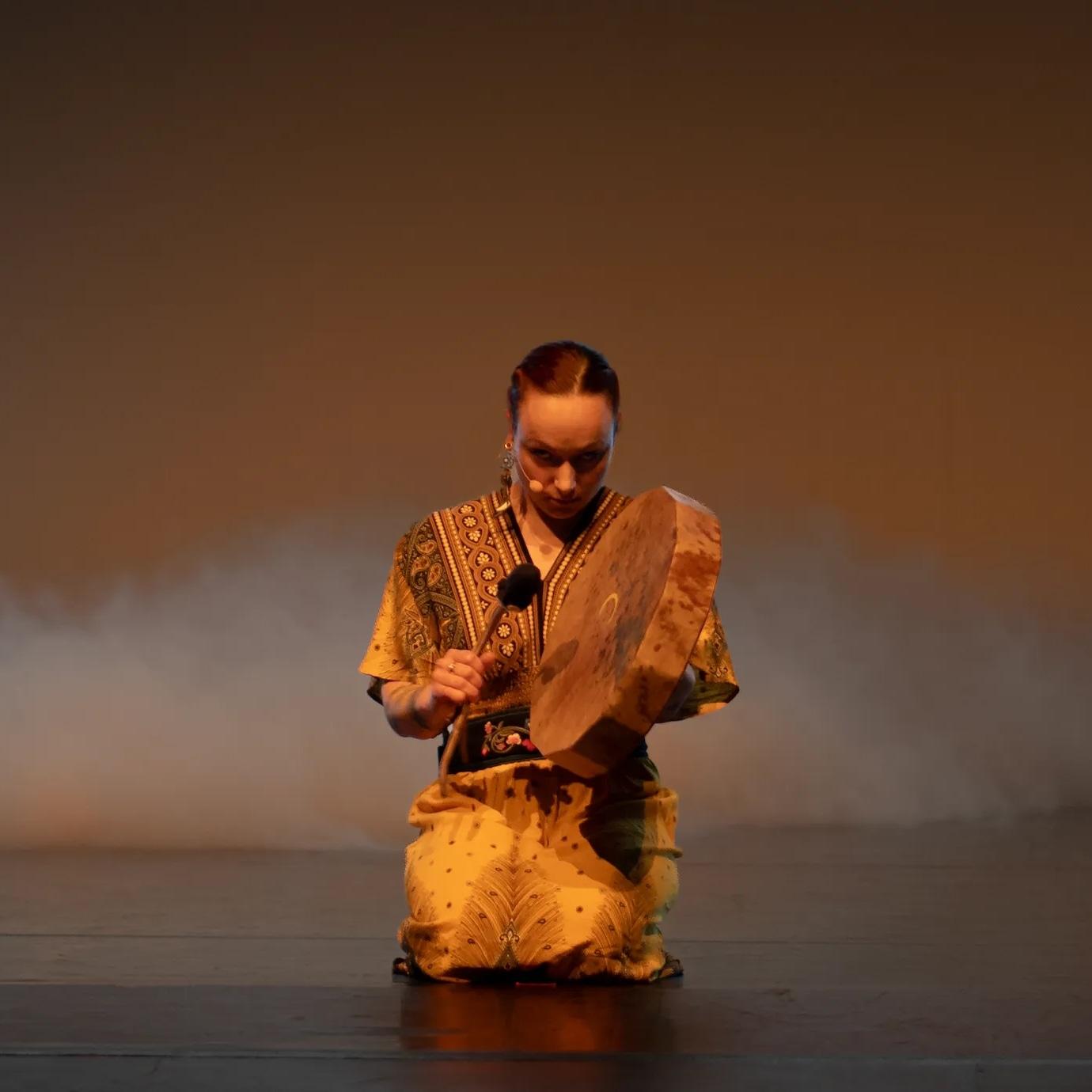 En danser i tradisjonell drakt spiller på en tromme på scenen, omgitt av tåke.