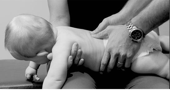 En baby får behandling på en benk, mens en voksen holder babyen og masserer ryggen.