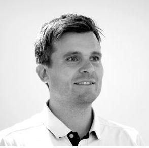 En smilende mann med kort, brunt hår iført en lys skjorte, står foran en lys bakgrunn.