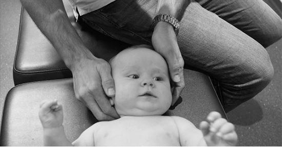 En voksen holder en babys hode under en behandling på en benk. Babyen ser nysgjerrig ut.