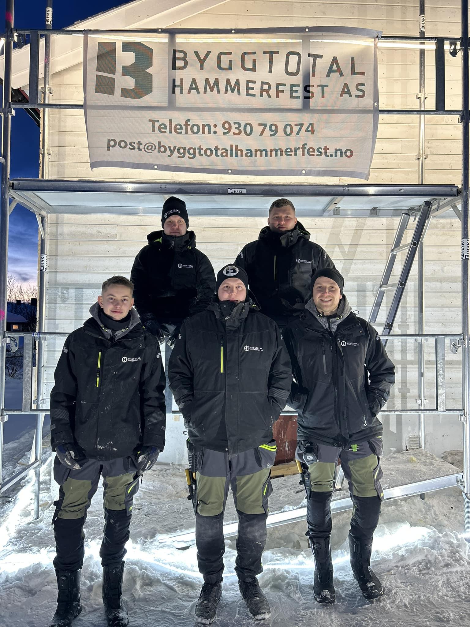 Fem arbeidere i arbeidsklær står foran en byggeplass med et skilt for Byggtotal Hammerfest AS.