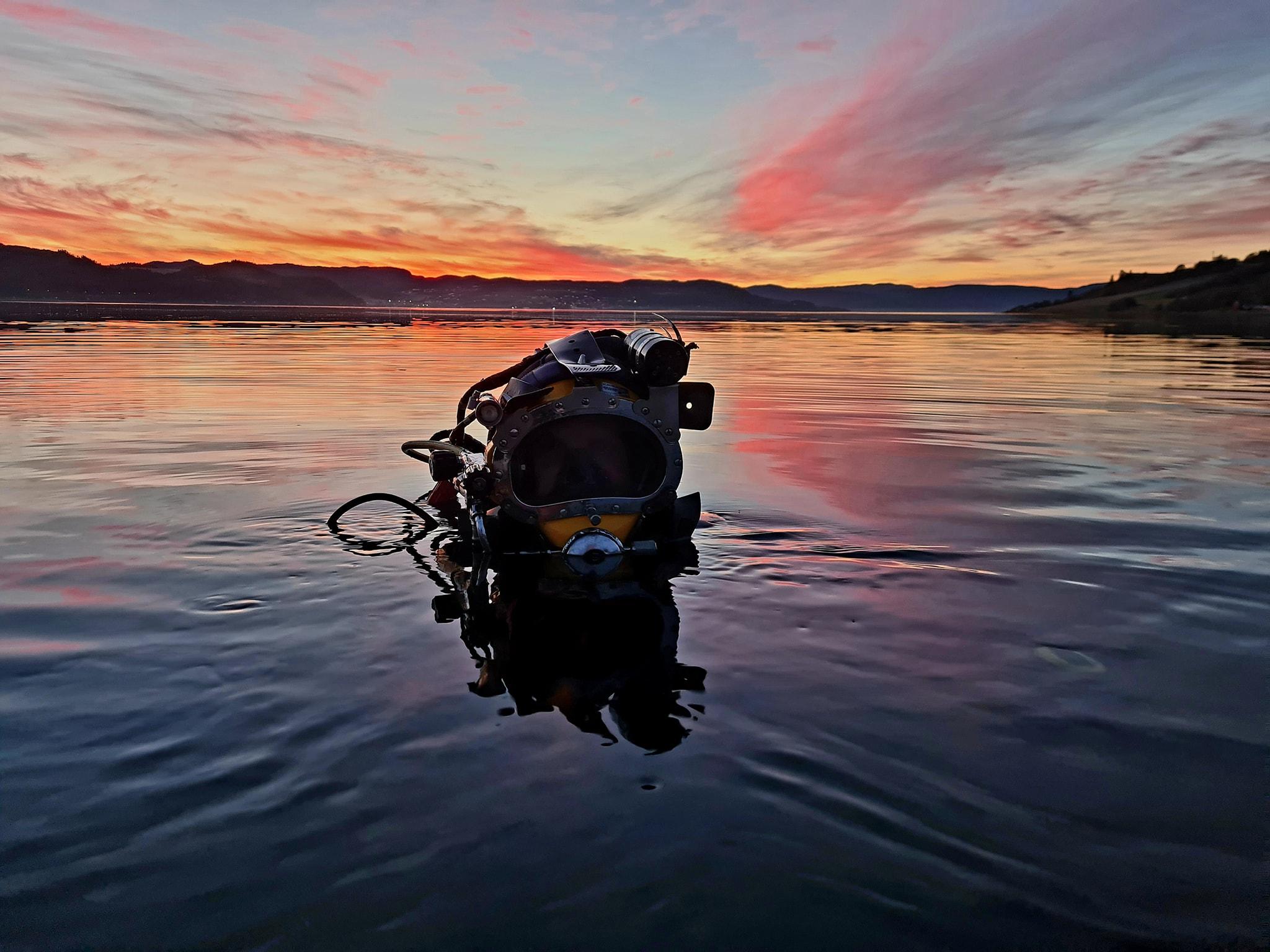 Dykker i vannet ved solnedgang, med fargerike skyer og speilbilde i overflaten.
