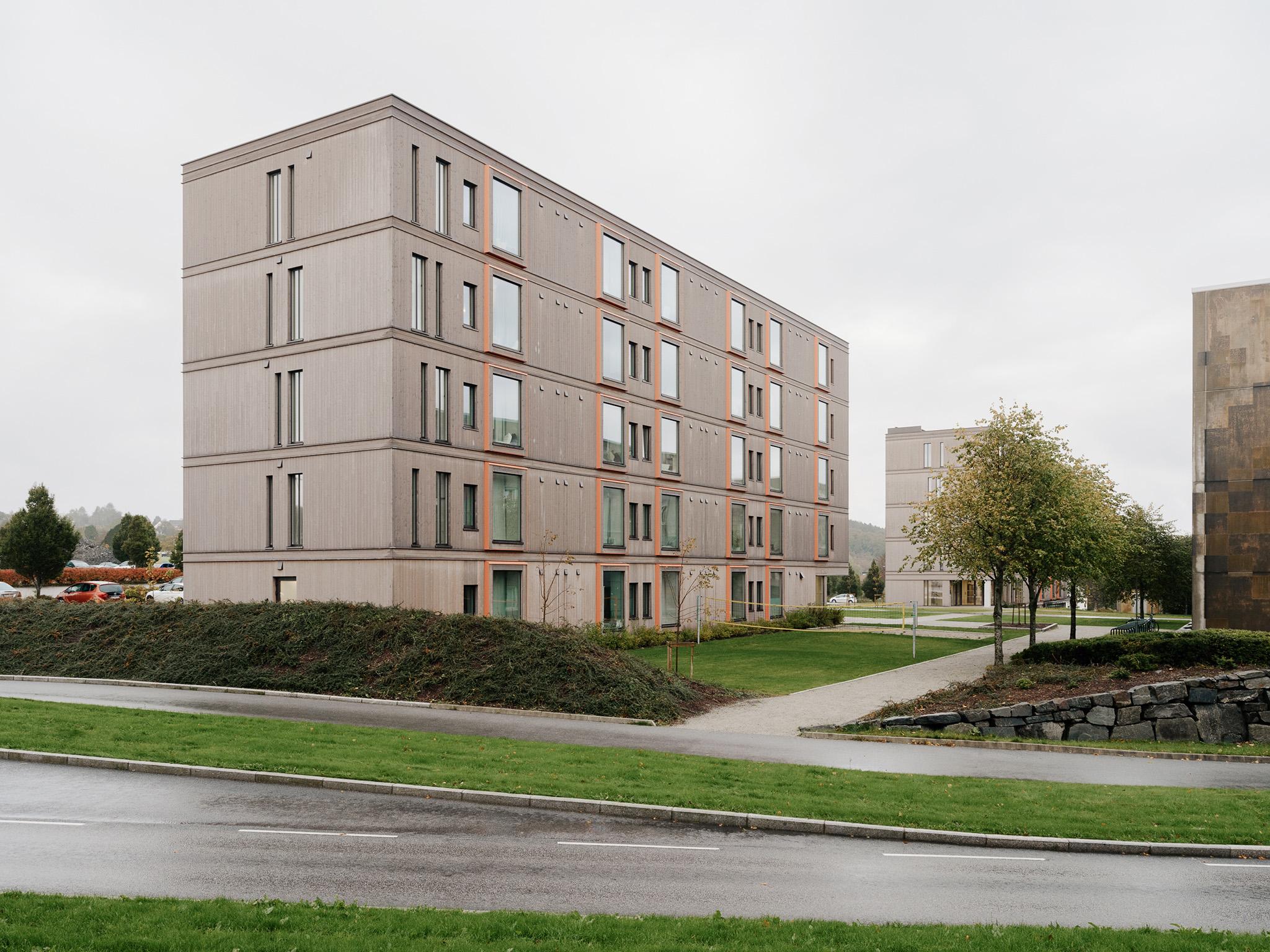 Moderne bygning med flere etasjer, store vinduer og grøntområde rundt, beliggende i et urbant miljø.