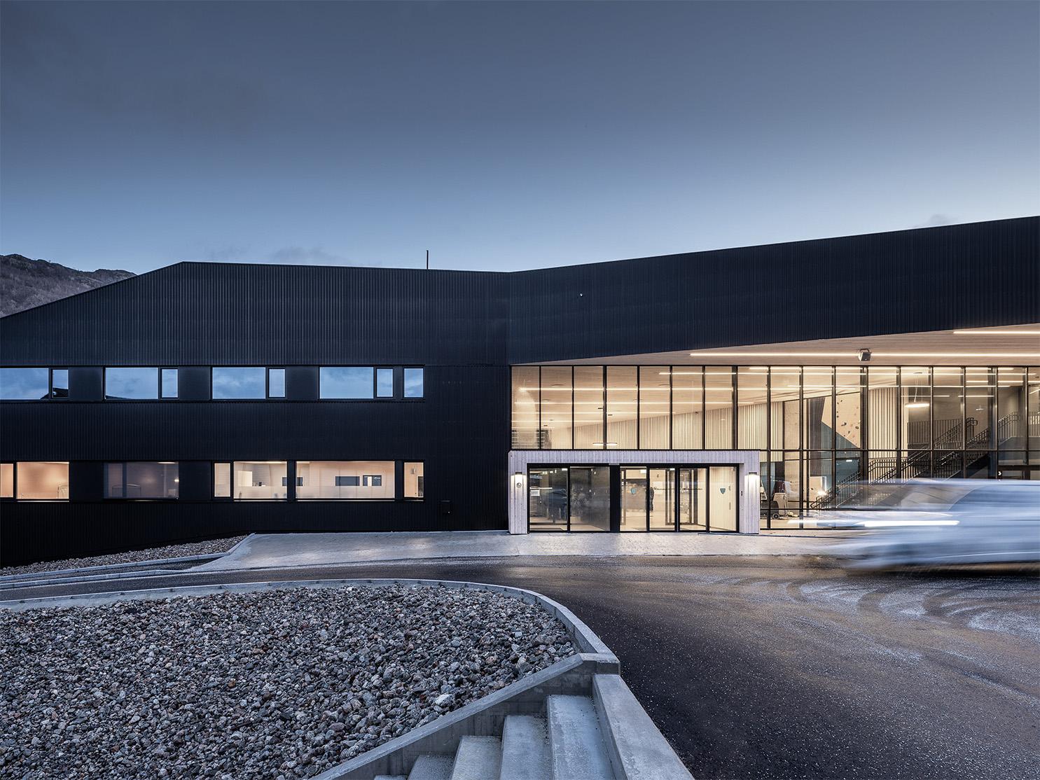 Moderne bygning med svart fasade og store vinduer, beliggende ved en grusvei i skumringen.