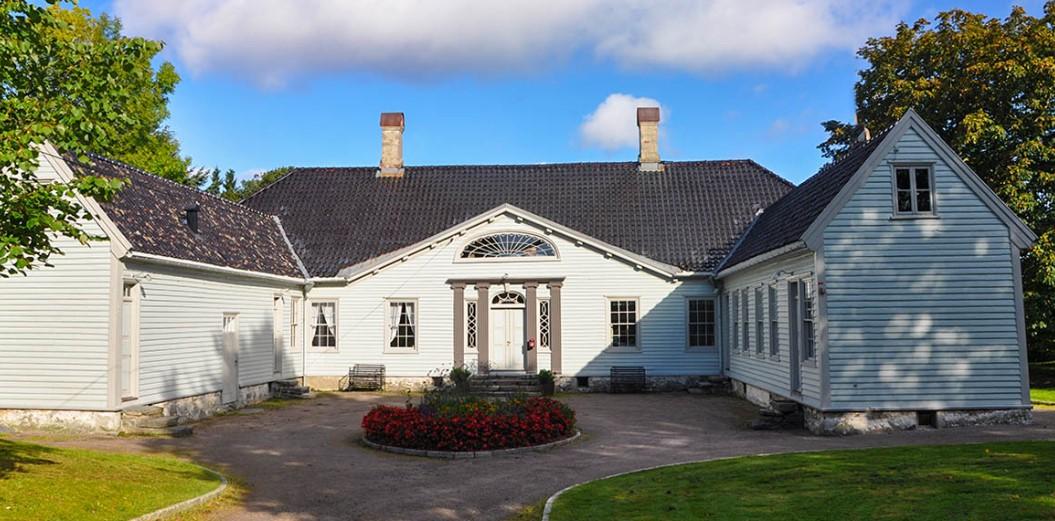 Et vakkert, blått hus med takvinduer og blomsterbed foran, omgitt av grønt landskap og blå himmel.