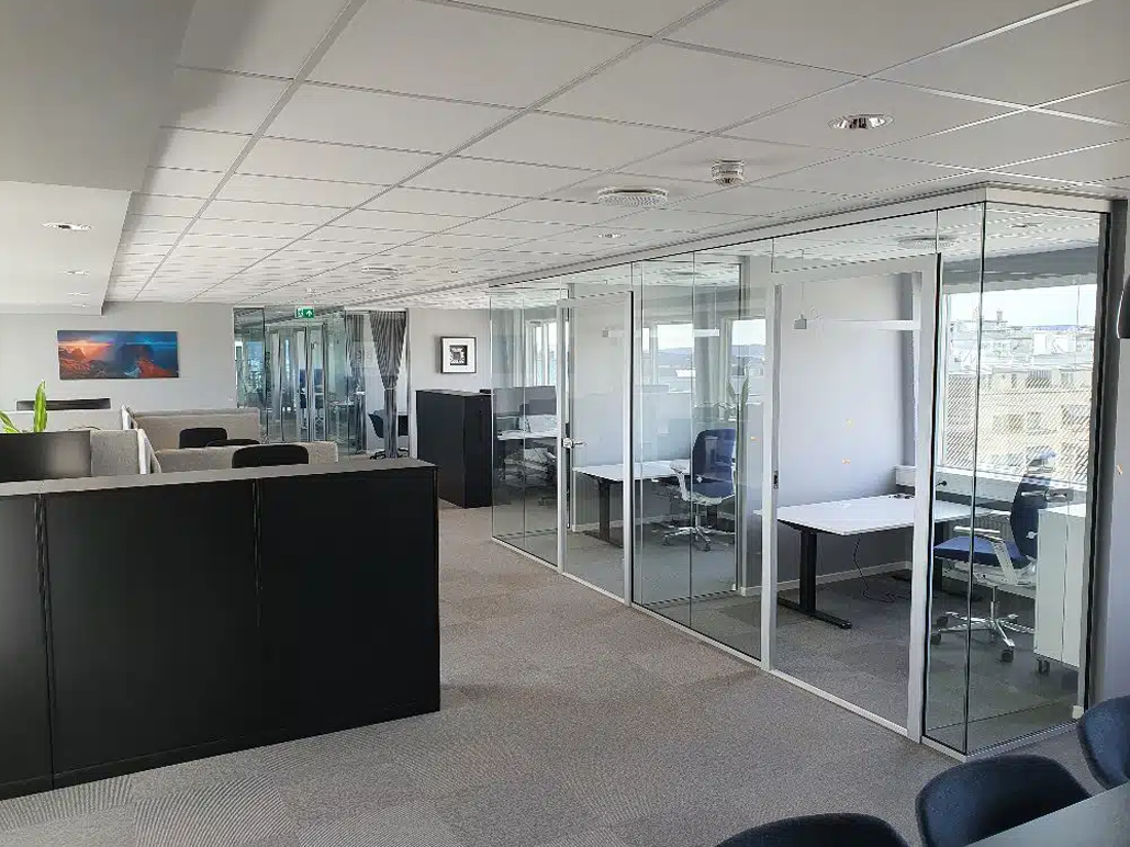 Moderne kontorlandskap med glassvegger, skrivebord og stoler, samt åpne arbeidsplasser.