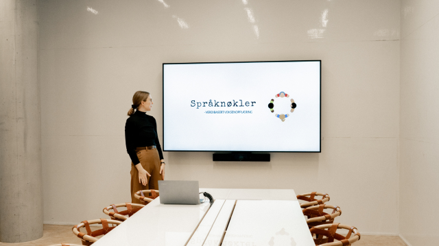 En kvinne står foran en stor skjerm i et møterom med logoen til Språknøkkel. Det er et moderne bord og stoler.