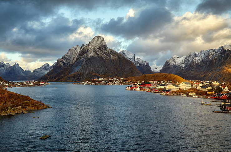 En vakker fjord med snødekte fjell og fargerike hus i en liten kystby under en dramatisk himmel.