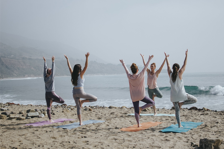 Fem kvinner praktiserer yoga på stranden med havet i bakgrunnen, iført komfortable treningsklær.