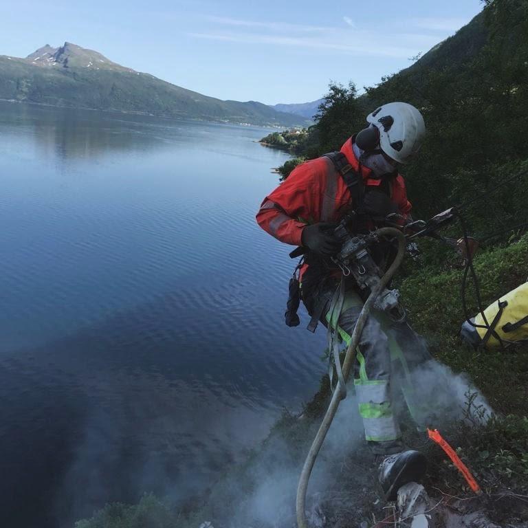 En person i redningsutstyr arbeider med en slange ved vannkanten, med fjell og innsjø i bakgrunnen.